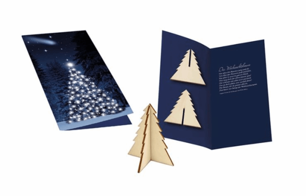 Eine personalisierte blaue Weihnachtskarte ist aufgeklappt. Sie beinhaltet einen kleinen hölzernen Tannenbaum, den man zusammenstecken kann.